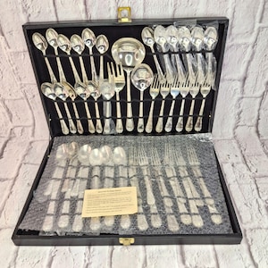 May include: A silver-plated cutlery set presented in a black case. The set includes spoons, forks, and knives, plus serving spoons and a ladle. The silverware features a decorative handle pattern and is neatly arranged for display.