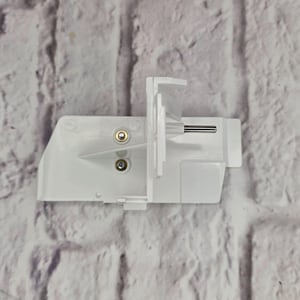 May include: A white plastic replacement part for a sewing machine, featuring a flat rectangular base with vertical support fins, two small circular metal contact points, and a single thin horizontal metal pin extending from the side.