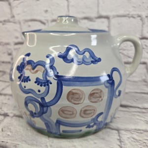 以下が含まれることがあります： 蓋付きのセラミックピッチャー。丸みを帯びたボディとハンドルが特徴です。ピッチャーは、白い背景に青と茶色の気まぐれなデザインで塗装されています。デザインには抽象的な形や模様が含まれており、フォークアートの美学を与えています。