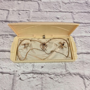 May include: A vintage jewelry set displayed in a cream-colored box. The set includes a necklace and earrings with gold-tone metal and dark red and clear accents, resting on a satin fabric.