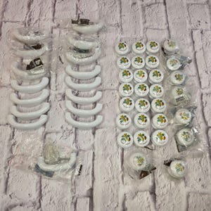 May include: A collection of white curved cabinet handles and round white cabinet knobs featuring a colorful floral pattern. The hardware pieces are individually wrapped in clear plastic packaging and arranged in rows on a textured white surface.