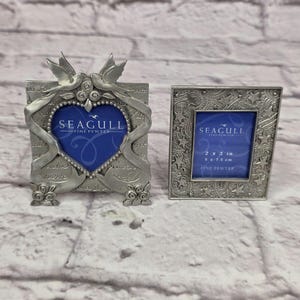 Puede incluir: Dos marcos de fotos plateados. El marco más grande tiene una abertura en forma de corazón con un fondo azul y el texto "SEAGULL FINE PEWTER". El marco más pequeño mide 5 x 7,5 cm y tiene un patrón de estrellas con el mismo texto.