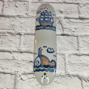 Puede incluir: Largo colgante de pared de cerámica blanquecina con temática náutica. Presenta un diseño de velero azul y naranja y un submarino, con olas y nubes azules. La pieza tiene dos agujeros para colgar.