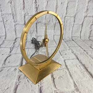 May include: A gold-toned metal electric clock featuring a circular frame with a transparent center and a single central hand. The clock is mounted on a rectangular base and includes a black electrical cord extending from the back of the mechanism.
