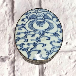 Puede incluir: Caja de baratijas ovalada con un diseño floral azul y blanco. La tapa presenta una gran flor central, rodeada de intrincadas vides y hojas azules sobre un fondo blanco. La caja tiene un borde de metal plateado.