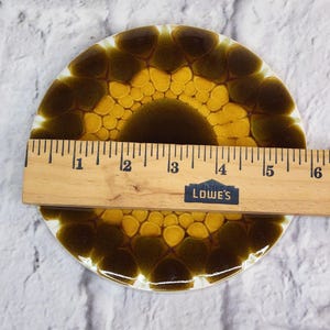 May include: A round, ceramic coaster with a sunflower design in shades of yellow and brown. A wooden ruler is placed across the coaster, measuring approximately 14 cm in diameter. The coaster has a white border.