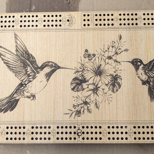 Könnte beinhalten: Cribbage-Spielbrett aus Holz mit einer detaillierten Gravur von zwei Kolibris und floralen Elementen. Das Brett hat eine ovale Form mit zahlreichen Löchern für die Stiftplatzierung und ist aus hellem Holz gefertigt.