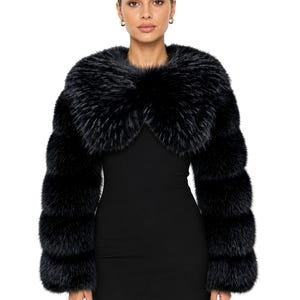 May include: A black faux fur bolero jacket featuring long sleeves and a wide, voluminous collar. The garment has a dense, textured appearance with horizontal paneling throughout the sleeves and body, worn over a simple black sleeveless dress.