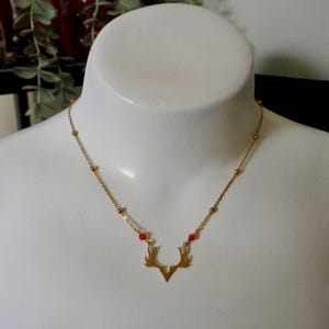May include: A gold-toned necklace featuring a central pendant shaped like stylised deer antlers. The delicate chain is accented with small gold beads and two tiny red beads positioned symmetrically on either side of the antler pendant.