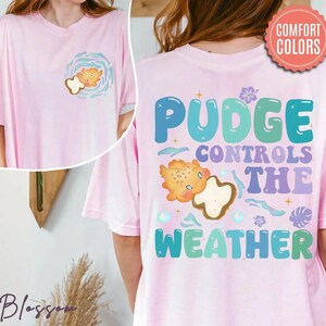 K&ouml;nnte beinhalten: Hellrosa T-Shirt mit einem Cartoon-Keks-Design. Auf der Vorderseite befindet sich eine Keks-Grafik in einem blauen Wirbel. Auf der R&uuml;ckseite des T-Shirts steht "PUDGE CONTROLS THE WEATHER" mit einer Keks-Grafik.