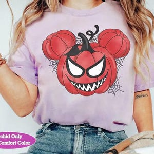 May include: A light purple short-sleeve t-shirt featuring a graphic of a red pumpkin with a sinister face and mouse-shaped ears, set against a black spiderweb background. The text "Orchid Only With Comfort Color" appears in a pink oval at the bottom.