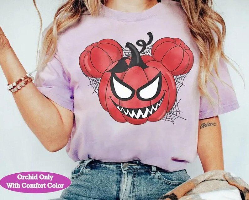 May include: A light purple short-sleeve t-shirt featuring a graphic of a red pumpkin with a sinister face and mouse-shaped ears, set against a black spiderweb background. The text "Orchid Only With Comfort Color" appears in a pink oval at the bottom.