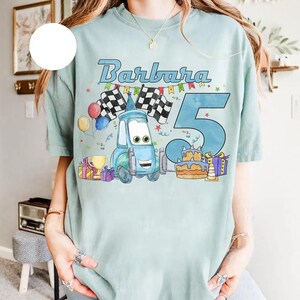 May include: Light blue t-shirt featuring the name "Barbara" and the number "5" in a playful font. The design includes a cartoon car, checkered flags, balloons, a birthday cake, and wrapped gifts, perfect for a birthday celebration.