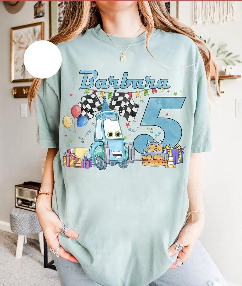 May include: Light blue t-shirt featuring the name "Barbara" and the number "5" in a playful font. The design includes a cartoon car, checkered flags, balloons, a birthday cake, and wrapped gifts, perfect for a birthday celebration.