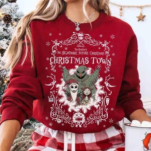May include: A red sweatshirt featuring characters from "The Nightmare Before Christmas." The design includes Jack Skellington, Sally, and Oogie Boogie, with the words "Christmas Town" in a decorative frame. The sweatshirt has long sleeves.