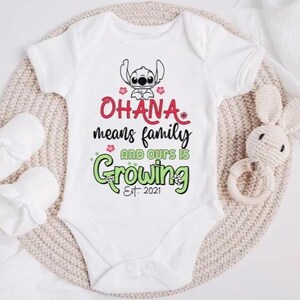 May include: White baby onesie with the text "OHANA means family AND OURS IS Growing Est. 2021" in red and green. A cartoon character is at the top. The onesie is on a beige knitted surface with a white stuffed bunny and baby mittens.