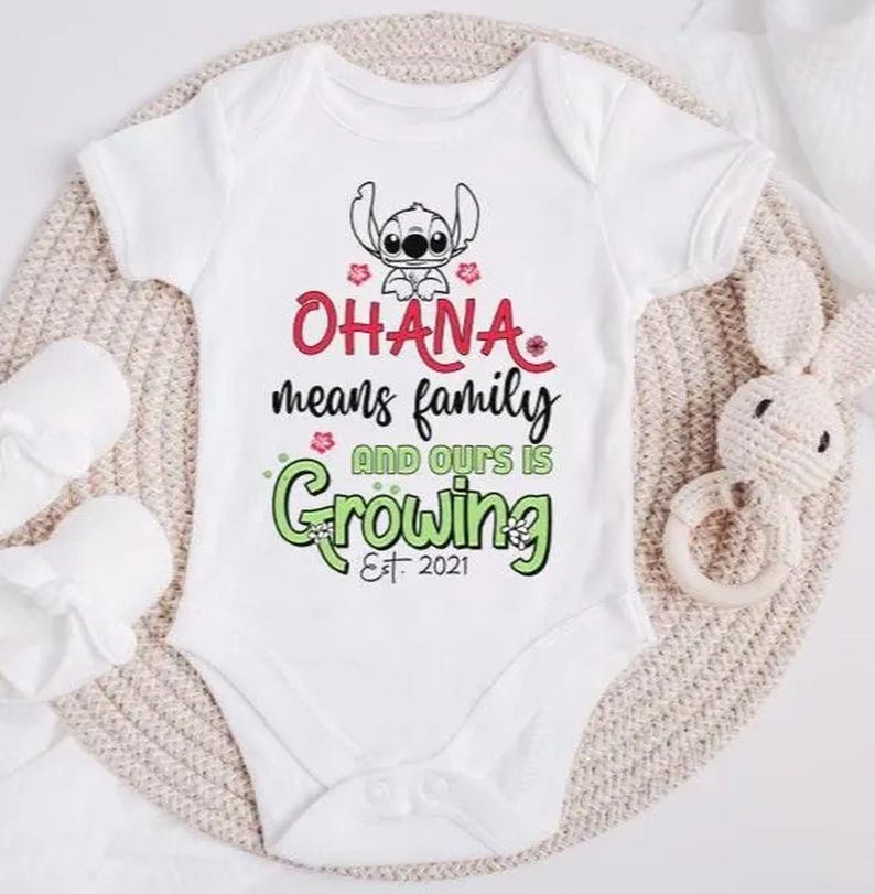 May include: White baby onesie with the text "OHANA means family AND OURS IS Growing Est. 2021" in red and green. A cartoon character is at the top. The onesie is on a beige knitted surface with a white stuffed bunny and baby mittens.