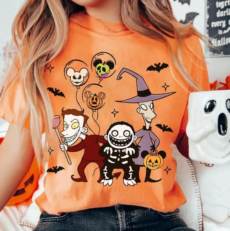 May include: Orange t-shirt with a Halloween cartoon graphic. The design features characters in costumes, bats, and Mickey Mouse-shaped balloons. The shirt is a casual style, suitable for the Halloween season.