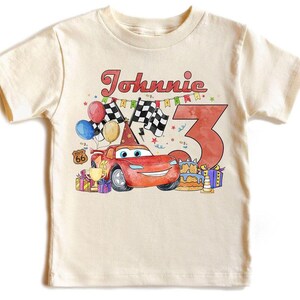 May include: A cream-colored t-shirt featuring a colorful graphic design. The design includes a red race car, the name "Johnnie" in red script, the number "3", balloons, checkered flags, and a birthday cake. The shirt is a children's apparel item.