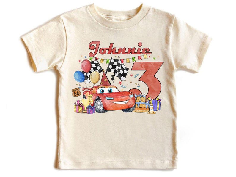 May include: A cream-colored t-shirt featuring a colorful graphic design. The design includes a red race car, the name "Johnnie" in red script, the number "3", balloons, checkered flags, and a birthday cake. The shirt is a children's apparel item.