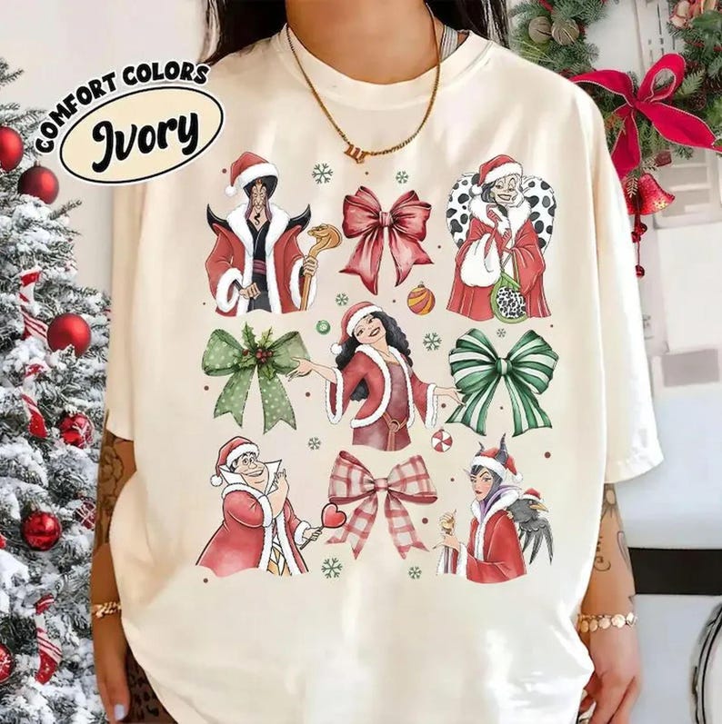 Op de afbeelding: Ivoor kleurig T-shirt met kerst-thema illustraties van schurken in kerstmutsen en strikken. Het shirt bevat de tekst "COMFORT COLORS" en "Ivory" in een ovaal. Het ontwerp bevat rode, groene en witte elementen.