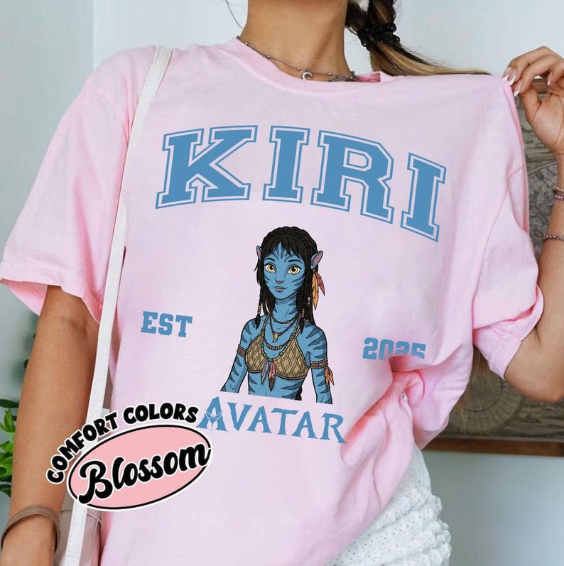 May include: Light pink t-shirt featuring the word "KIRI" in blue block letters above an illustration of a blue Avatar character. The shirt also includes the words "EST" and "2025", and the word "AVATAR" below the illustration. A "Comfort Colors Blossom" logo is on the bottom left.