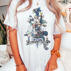 Puede incluir: Camiseta blanca con un gr&aacute;fico colorido de personajes de Kingdom Hearts, incluyendo a Mickey Mouse y el Pato Donald. El dise&ntilde;o muestra varias figuras del juego, con el logo de 'Kingdom Hearts' en la parte inferior.