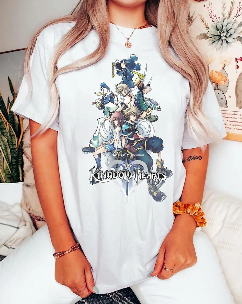 Puede incluir: Camiseta blanca con un gr&aacute;fico colorido de personajes de Kingdom Hearts, incluyendo a Mickey Mouse y el Pato Donald. El dise&ntilde;o muestra varias figuras del juego, con el logo de 'Kingdom Hearts' en la parte inferior.