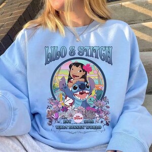 Może przedstawiać: Jasnoniebieska bluza z napisem "Lilo & Stitch" i ilustracją Lilo i Stitch otoczonych kwiatami. Widoczny jest r&oacute;wnież tekst "EST 2002 Walt Disney World".