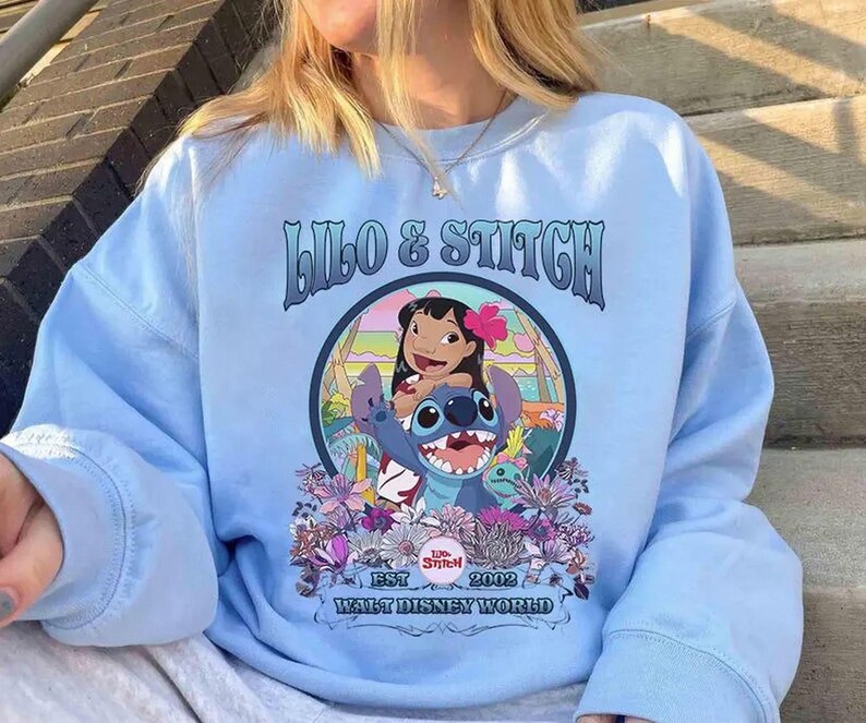 Może przedstawiać: Jasnoniebieska bluza z napisem "Lilo & Stitch" i ilustracją Lilo i Stitch otoczonych kwiatami. Widoczny jest r&oacute;wnież tekst "EST 2002 Walt Disney World".
