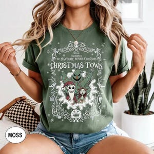 May include: Moss green t-shirt featuring a graphic from "The Nightmare Before Christmas." The design includes Jack Skellington, Sally, and Oogie Boogie, with the text "Christmas Town" in a decorative frame. The shirt has short sleeves.