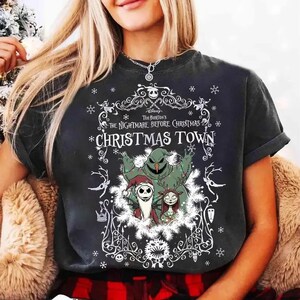 May include: Dark gray t-shirt featuring a graphic from Tim Burton's "The Nightmare Before Christmas." The design includes Jack Skellington, Sally, and Oogie Boogie, with the words "Christmas Town" in a decorative frame. Snowflakes and decorative elements surround the characters.