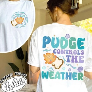 K&ouml;nnte beinhalten: Wei&szlig;es Comfort Color T-Shirt mit Grafik auf Vorder- und R&uuml;ckseite. Die Grafik auf der Vorderseite zeigt eine Cartoon-Kreatur in einem Wirbel. Die Grafik auf der R&uuml;ckseite zeigt "Pudge Controls The Weather" mit einer passenden Cartoon-Kreatur.