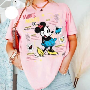 以下が含まれることがあります： 水玉模様の青いドレスと黄色の靴を履いたミニーマウスが描かれたピンクのTシャツ。「MINNIE MOUSE」の文字と「Exhibit B: The Style Icon」などのグラフィック要素が含まれています。
