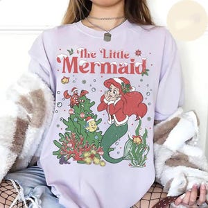 K&ouml;nnte beinhalten: Ein helllila Langarmshirt mit einem weihnachtlichen Motiv von Arielle, der Meerjungfrau. Das Design zeigt einen Weihnachtsbaum, Fabius, Sebastian und den Text "The Little Mermaid" in Rot.