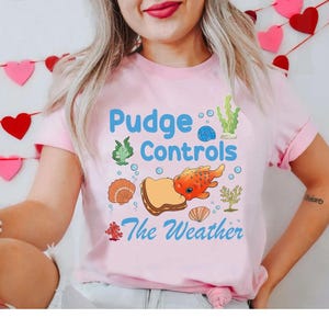 May include: A light pink t-shirt with the text "Pudge Controls The Weather" in blue and a cartoon graphic of a fish, bread, and seashells. The shirt has short sleeves.