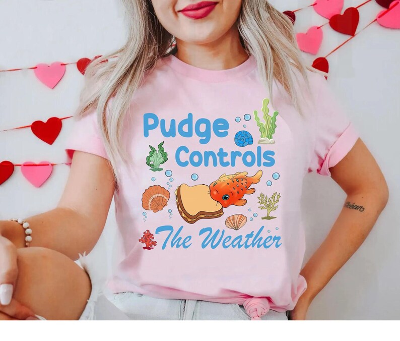 May include: A light pink t-shirt with the text "Pudge Controls The Weather" in blue and a cartoon graphic of a fish, bread, and seashells. The shirt has short sleeves.