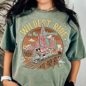 以下が含まれることがあります： セージグリーンの半袖Tシャツ。線路を走る列車と漫画のキャラクターが描かれています。黄色と青の文字で「WILDEST RIDE in the wilderness」と書かれています。デザインにはピンクの山々と茶色の円形の縁取りが含まれています。