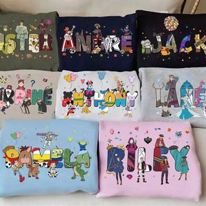 May include: A selection of sweatshirts in different colours, each displaying a name in vibrant, cartoon lettering. The names are complemented by illustrations of animated characters, resulting in a personalised and playful design.