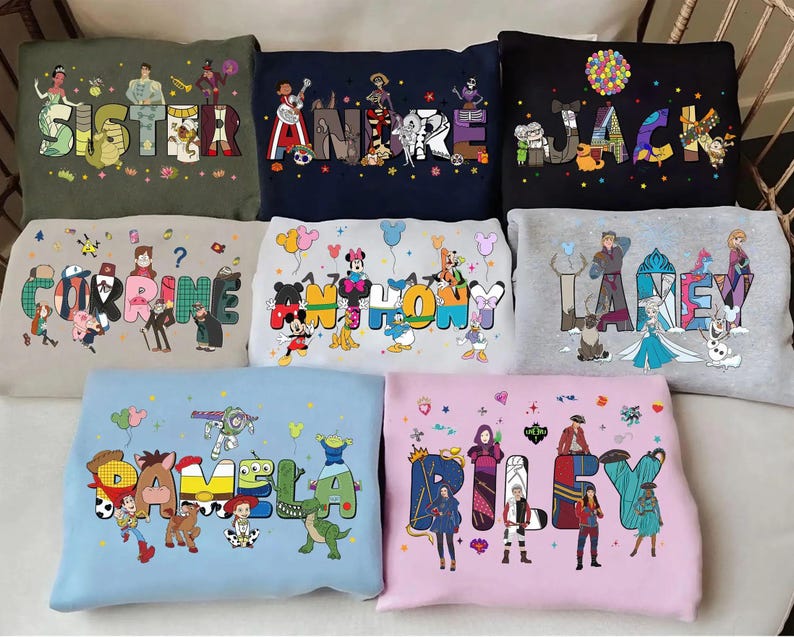 May include: A selection of sweatshirts in different colours, each displaying a name in vibrant, cartoon lettering. The names are complemented by illustrations of animated characters, resulting in a personalised and playful design.