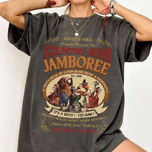 May include: Dark gray t-shirt featuring the text "Grizzly Hall Proudly Presents The Country Bear Jamboree" in red and orange. The shirt has a vintage design with a cartoon band of bears playing instruments. The text "Est. 1898" is also visible.