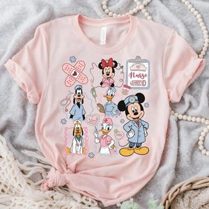 Puede incluir: Una camiseta rosa claro con personajes de dibujos animados vestidos de enfermeras. El dise&ntilde;o incluye a Minnie Mouse, Mickey Mouse, el Pato Donald, Goofy y Daisy, junto con gr&aacute;ficos m&eacute;dicos como vendas y una carpeta.