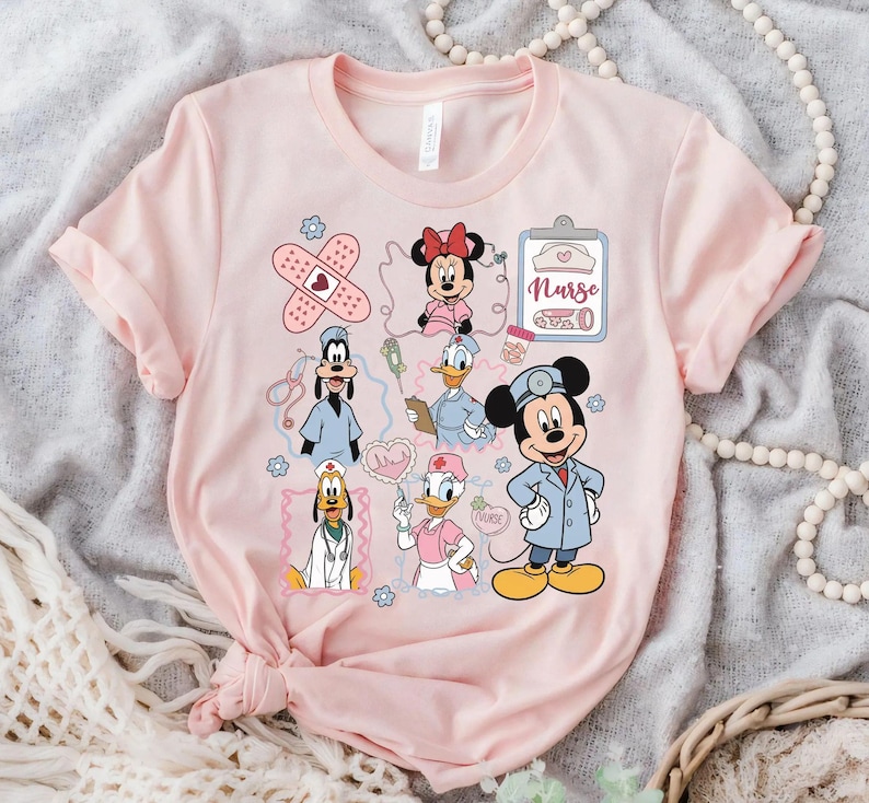 Puede incluir: Una camiseta rosa claro con personajes de dibujos animados vestidos de enfermeras. El dise&ntilde;o incluye a Minnie Mouse, Mickey Mouse, el Pato Donald, Goofy y Daisy, junto con gr&aacute;ficos m&eacute;dicos como vendas y una carpeta.