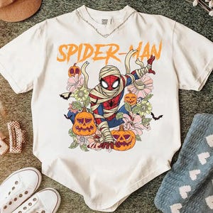 K&ouml;nnte beinhalten: Wei&szlig;es T-Shirt mit einem Spider-Man-Motiv als Mumie, umgeben von K&uuml;rbissen und Blumen. Der orangefarbene Text "SPIDER-MAN" steht dar&uuml;ber. Das T-Shirt hat kurze &Auml;rmel und einen Rundhalsausschnitt.