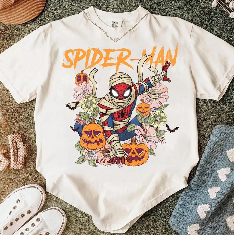 K&ouml;nnte beinhalten: Wei&szlig;es T-Shirt mit einem Spider-Man-Motiv als Mumie, umgeben von K&uuml;rbissen und Blumen. Der orangefarbene Text "SPIDER-MAN" steht dar&uuml;ber. Das T-Shirt hat kurze &Auml;rmel und einen Rundhalsausschnitt.