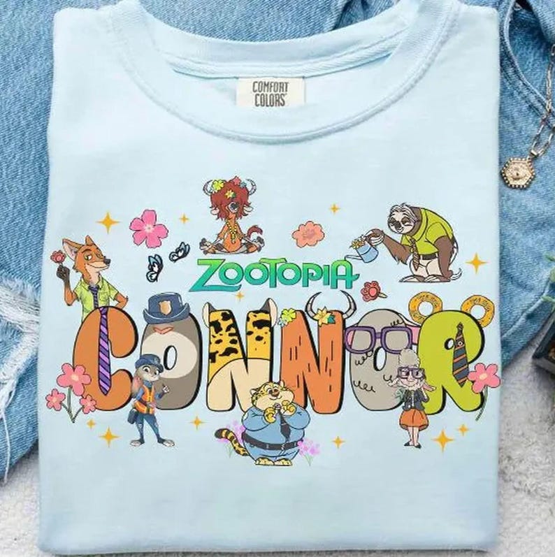 Puede incluir: Camiseta Comfort Colors azul claro con la palabra "Zootopia" y el nombre "Connor" en un dise&ntilde;o colorido y de dibujos animados. El dise&ntilde;o incluye varios personajes de animales y acentos florales, creando una est&eacute;tica l&uacute;dica y vibrante.