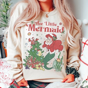 K&ouml;nnte beinhalten: Beiges Sweatshirt mit einem festlichen Aufdruck von Arielle, der Meerjungfrau. Das Design zeigt Arielle in einem roten Mantel und gr&uuml;nem Schwanz, einen Weihnachtsbaum und den Text "The Little Mermaid" in Rot und Wei&szlig;.