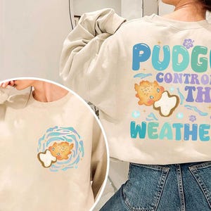 K&ouml;nnte beinhalten: Beigefarbenes Sweatshirt mit dem Text "Pudge Controls The Weather" in Blau, Gr&uuml;n und Lila. Das Design beinhaltet eine Cartoonfigur und Wettergrafiken. Eine kleinere Version des Designs befindet sich auf der Vorderseite.