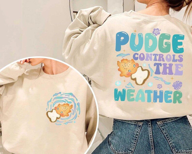 K&ouml;nnte beinhalten: Beigefarbenes Sweatshirt mit dem Text "Pudge Controls The Weather" in Blau, Gr&uuml;n und Lila. Das Design beinhaltet eine Cartoonfigur und Wettergrafiken. Eine kleinere Version des Designs befindet sich auf der Vorderseite.