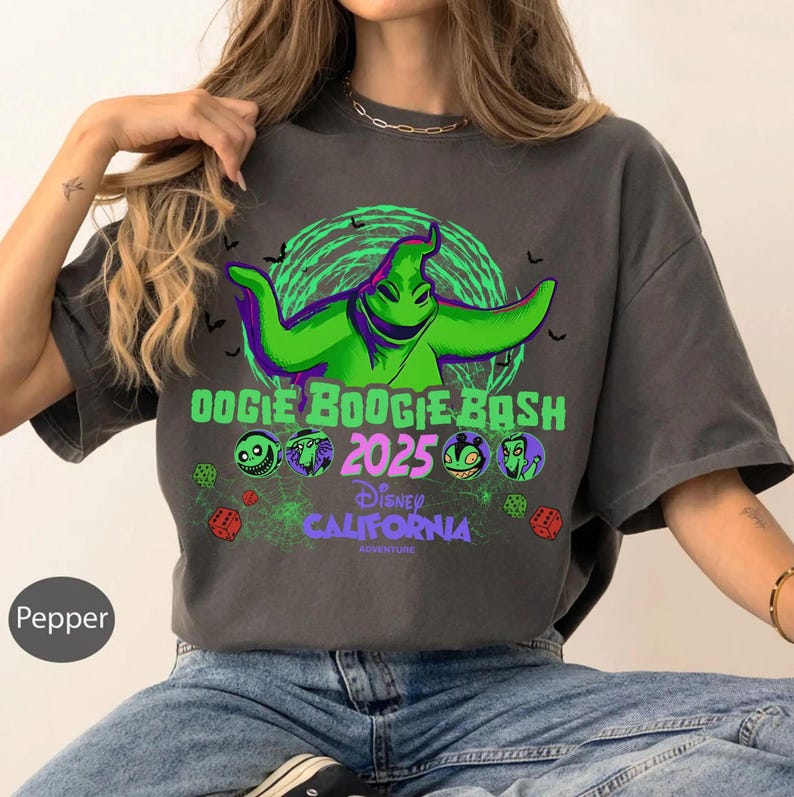 Op de afbeelding: Een antracietgrijs t-shirt met een grote groene afbeelding van het personage Oogie Boogie, de tekst Oogie Boogie Bash 2025, Disney California Adventure, en kleine ronde karakterportretten, rode dobbelstenen en spinnenwebpatronen.